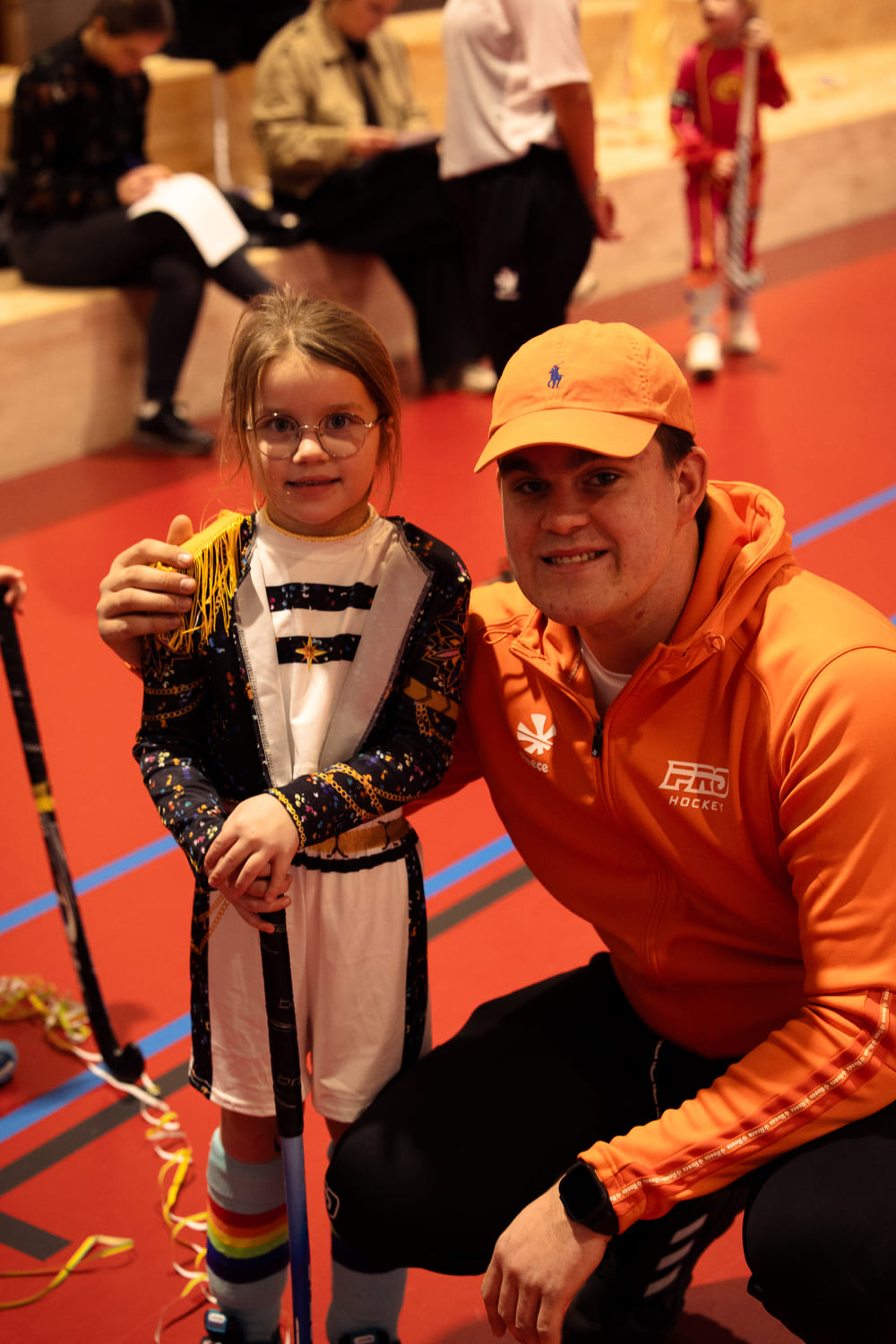 Kind in kleurrijke hockeykleding poseert met een coach in oranje Pro Hockey-kleding tijdens een hockeikamp in een sporthal.