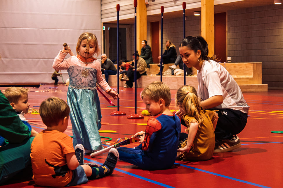 Kinderen in kleurrijke kostuums nemen deel aan een ProHockey kampactiviteit in een sporthal, bijgestaan door een begeleider.