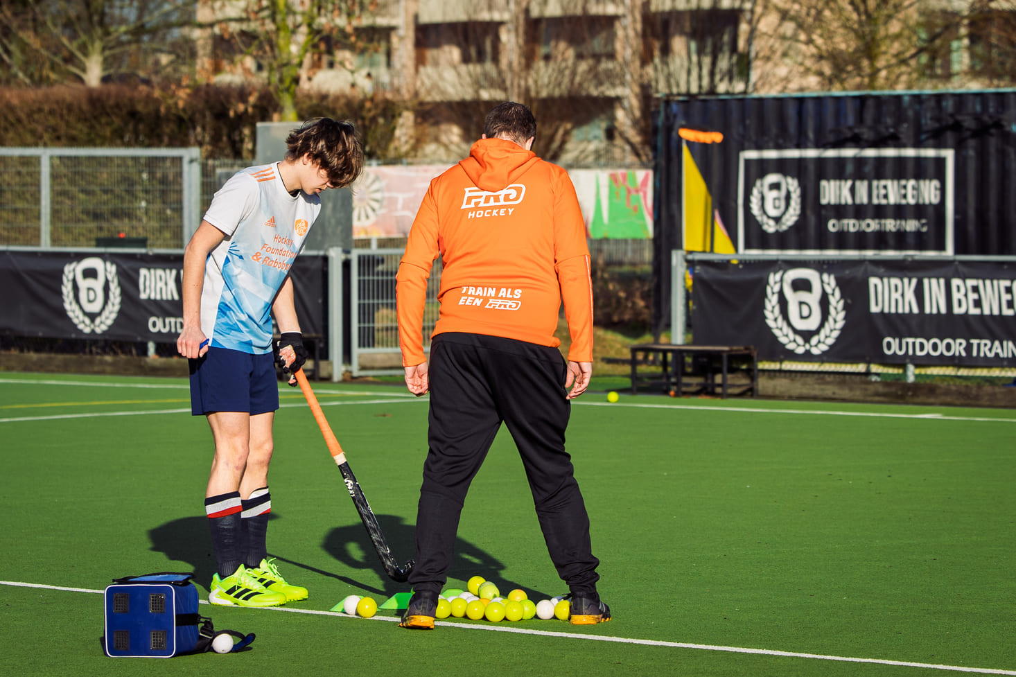 Twee personen tijdens een hockeytraining op een veld, waarbij de coach van ProHockey tips geeft aan een jonge speler, omringd door felgekleurde hockeyballen tijdens een hockeekamp.