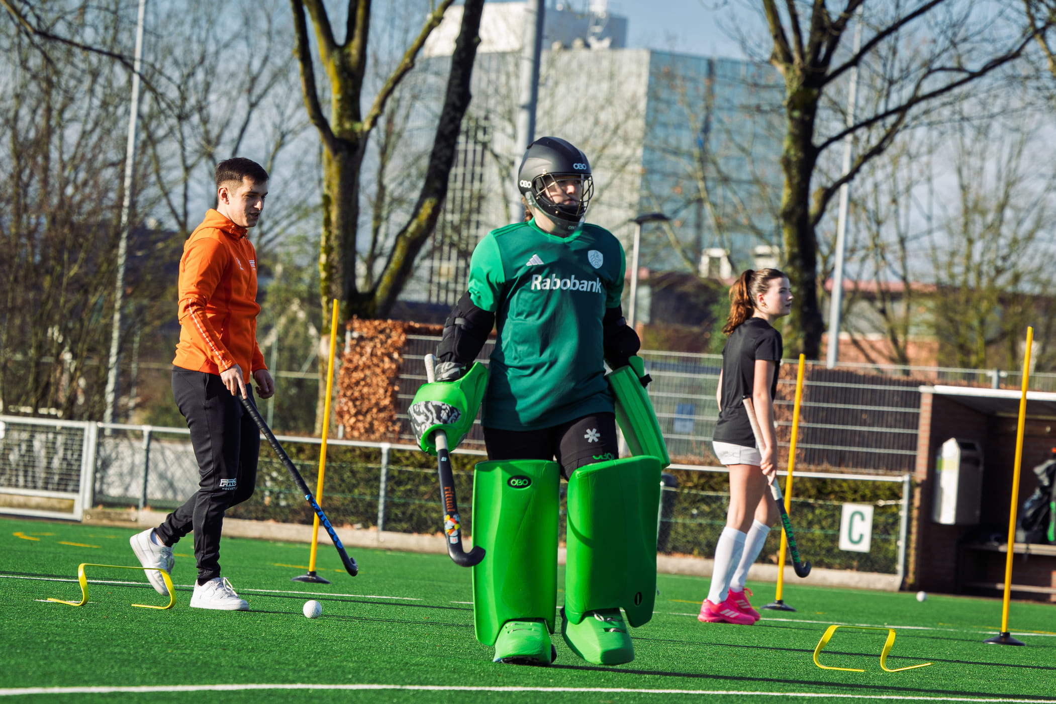 Afbeelding van een hockeykeeper in een groen tenue tijdens een ProHockey training op een veld, omringd door andere spelers en gele hindernissen, ideaal voor hockeykampen.