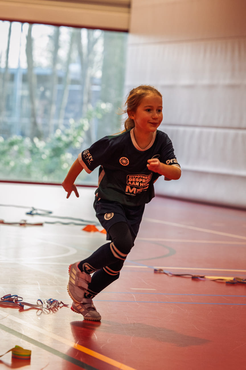 Jong meisje rent enthousiast tijdens een ProHockey training in een sporthal met kleurrijke hockey accessoires op de achtergrond.