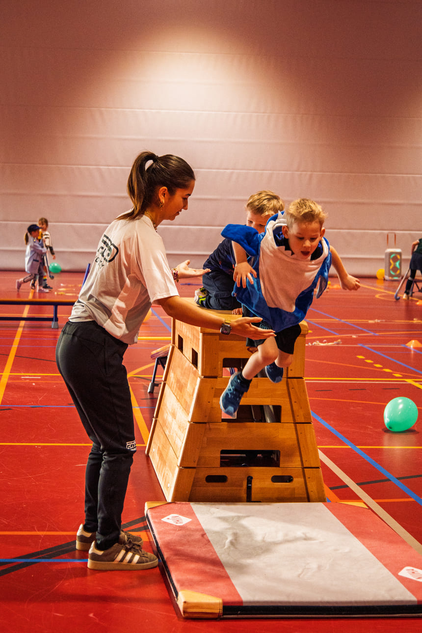 Kind springt over kast in sportzaal onder begeleiding van coach tijdens ProHockey hockeykampen.