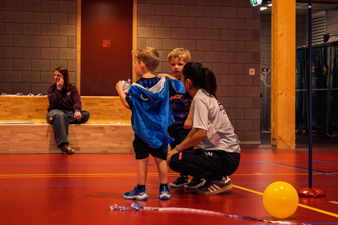 Kinderen en coach tijdens een activiteit in een sporthal, met een blauwe dierencape en een gele ballon.