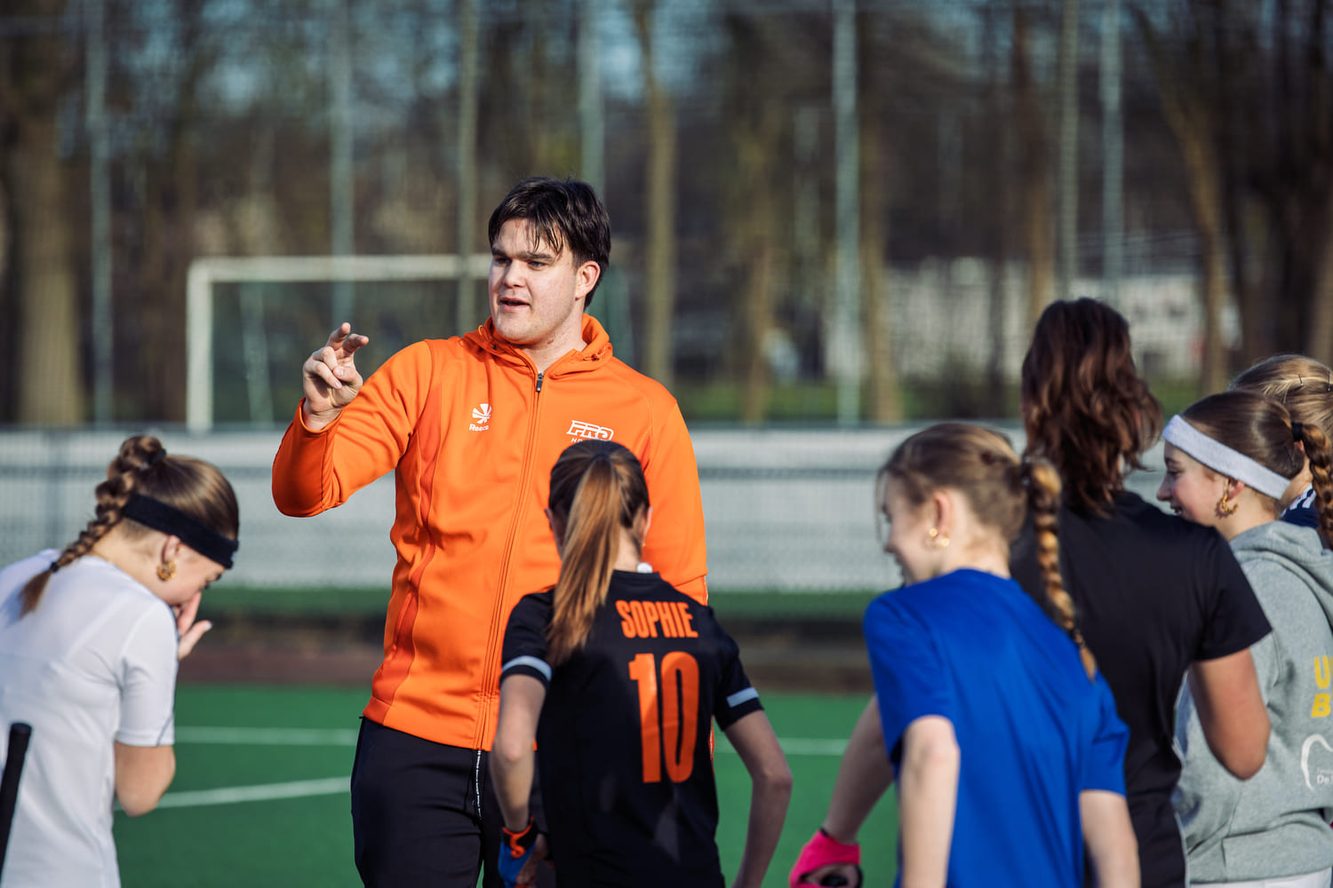 Coach in oranje ProHockey-jack geeft hockeytraining aan jonge spelers op veld.