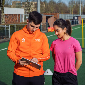 Twee personen op een hockeyveld tijdens een ProHockey training, waarbij een van hen een tablet vasthoudt. Context: hockekampen.