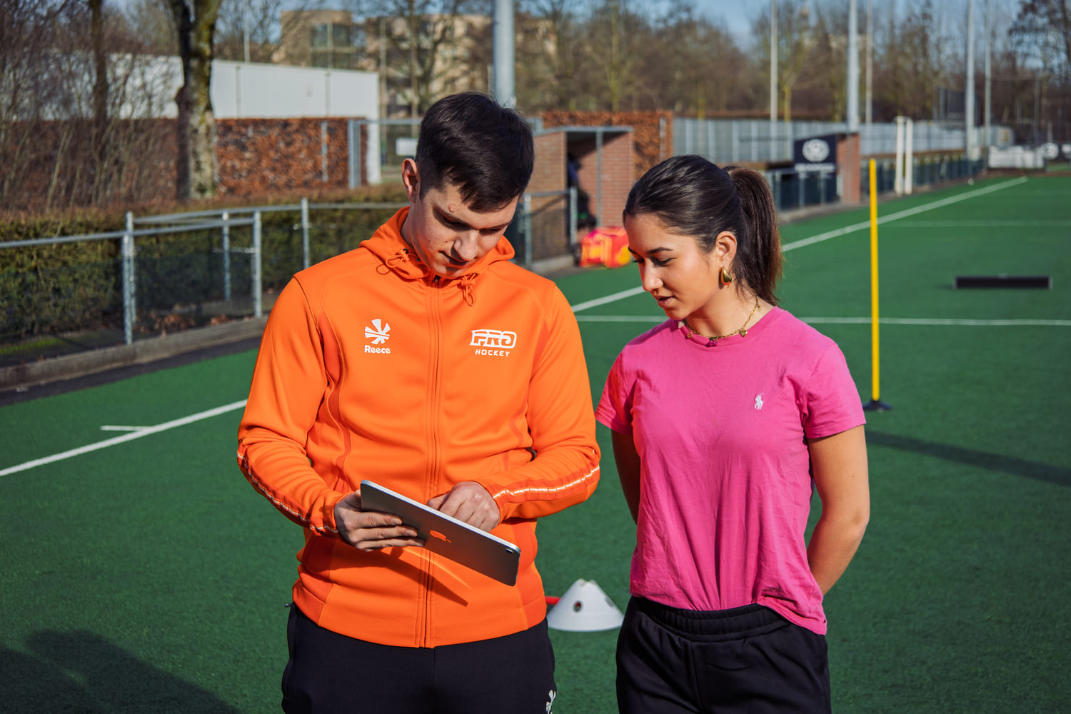 Twee personen op een hockeyveld tijdens een ProHockey training, waarbij een van hen een tablet vasthoudt. Context: hockekampen.