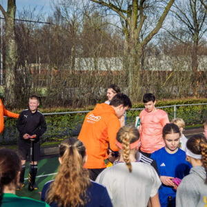 Groep jonge hockeyspelers en trainers verzamelen zich op een veld tijdens een ProHockey-kamp, gekleed in sportieve oranje en blauwe outfits.