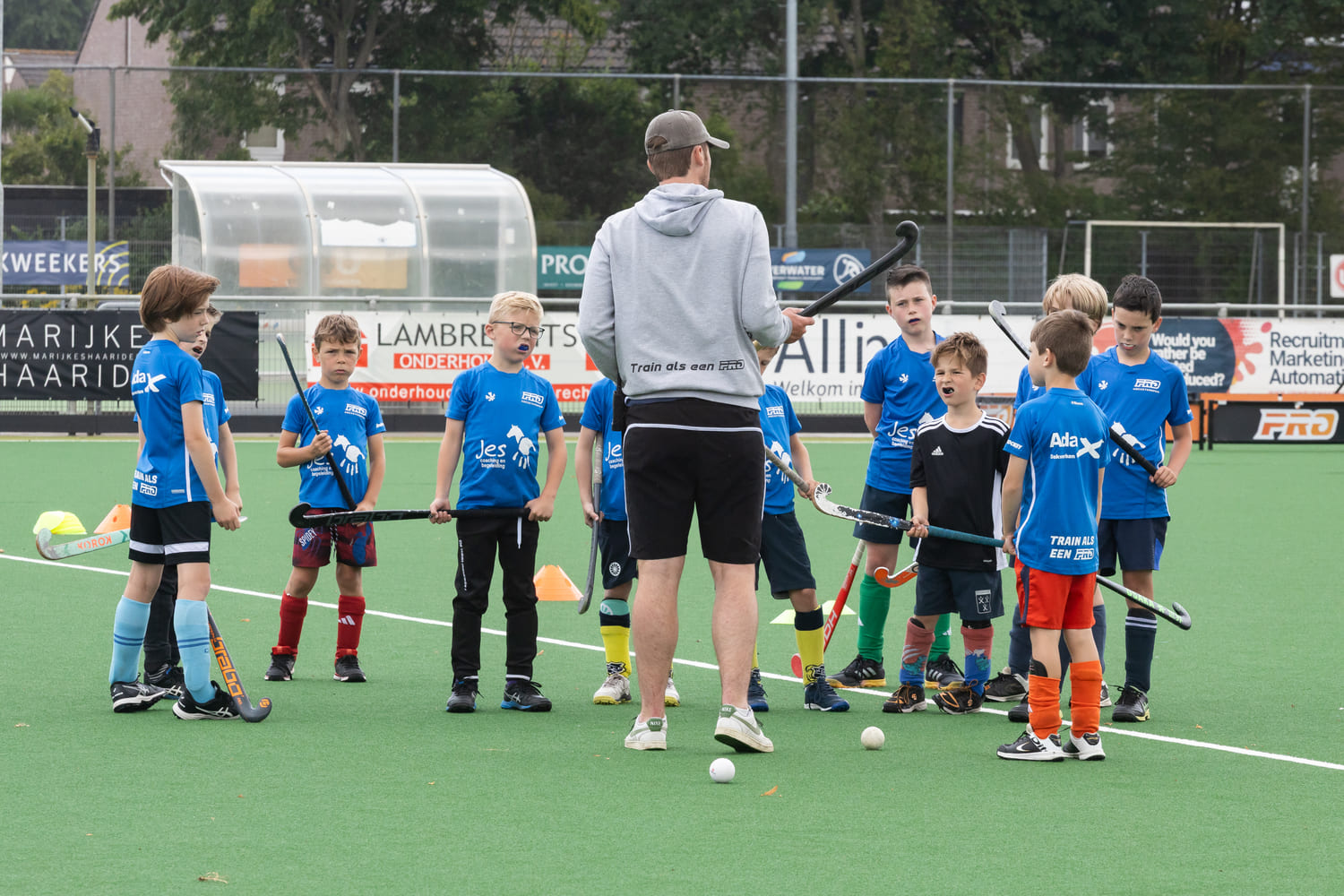Kinderen tijdens een hockeytraining op ProHockey zomerkamp bij hockeyclub Prinsenbeek, begeleid door een trainer die instructies geeft op het groene veld.